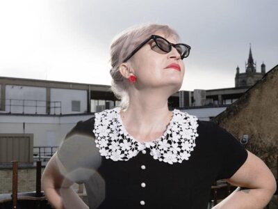 photo of Helen Ivory looking up at the sky wearing a black top with a little lacy white collar, red earrings, red lippy and black sunglasses, against buildings. Definitely a foxy rockabilly vibe going on here.