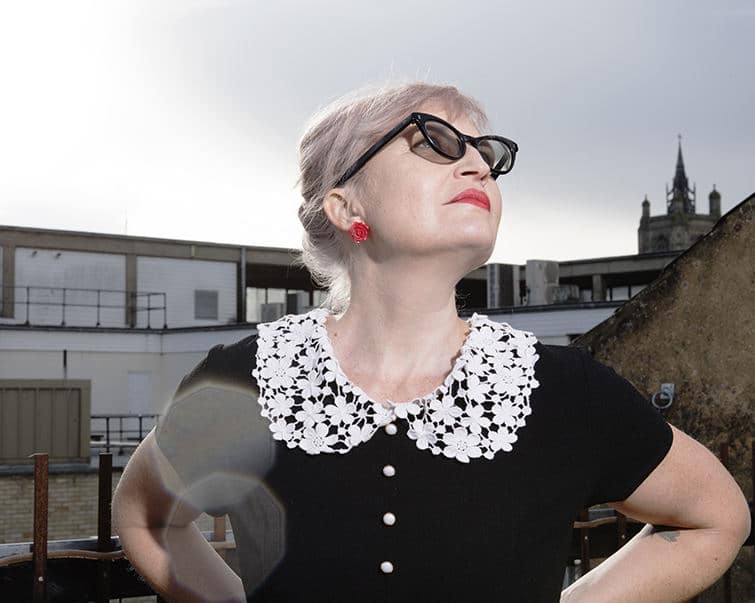 photo of Helen Ivory looking up at the sky wearing a black top with a little lacy white collar, red earrings, red lippy and black sunglasses, against buildings. Definitely a foxy rockabilly vibe going on here.