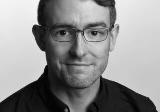 Black and white photo of Ben Wilkinson, a young friendly looking man with glasses