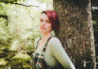 Kim Moore with red hair wearing a green top and green check dungarees leaning against a tree