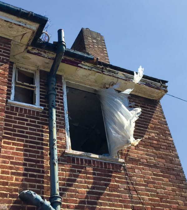 A brick built house with a window opening that has no window and a billowing white plastic curtain, there is a blue sky and an air of abandonment.