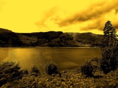 A lake, with a hill behind tinted yellow, kind of sepia but are yellow. The picture has been taken from a bus and a faint reflection of the photographer can be barely seen