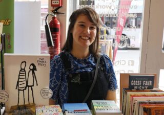 Picture of Emma smiling with shoulder length brown hair wearing blue shirt and dungarees sitting behind a table full of books.