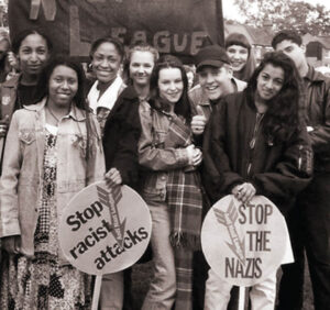 Black and white image of young people at an anti Nazi rally, they are carrying banners with "stop the nazis" and "stop racist attacks"