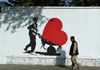 Photograph of a man walking by a white wall in Kabul, on the wall is an illustration of a man pushing a wheelbarrow with a large red heart shape in it