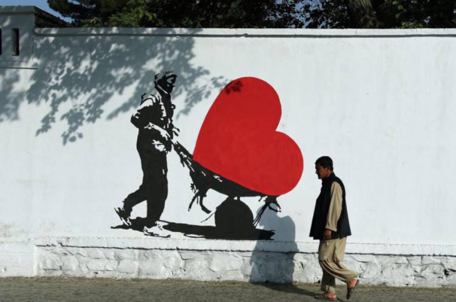 Photograph of a man walking by a white wall in Kabul, on the wall is an illustration of a man pushing a wheelbarrow with a large red heart shape in it