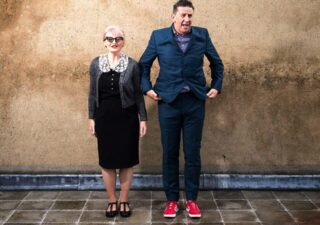 Martin and Helen standing in front of a wall, he is wearing a blue suit with red trainers, she is wearing a slightly retro frock in black with a white lace collar, she has fifty style glasses and a grey cardigan.