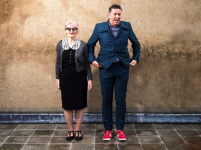 Martin and Helen standing in front of a wall, he is wearing a blue suit with red trainers, she is wearing a slightly retro frock in black with a white lace collar, she has fifty style glasses and a grey cardigan.
