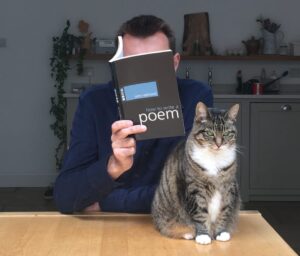 a man sitting at a table reading a book which hides his face, also on the table is a tabby cat