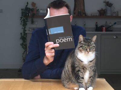 a man sitting at a table reading a book which hides his face, also on the table is a tabby cat