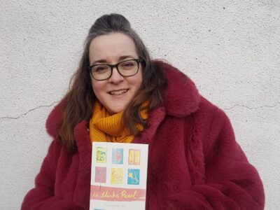 Julia Bird holding her book, she has medium length brown hair and is wearing a purple faux fur coat with a yellow wool scarf. she is standing in front of a white wall.