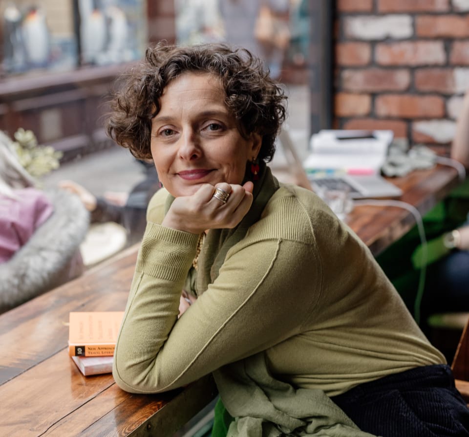 Sophie sitting at a desk turning to look at the camera, she has a green jumper, two books are on the desk, her chin is resting on her hand.