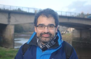 Photo of Tom standing on a a river bank with a bridge in the background. He has a dark beard and glasses and is wearing a blue waterproof jacket and a grey check scarf