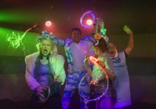 Four people dressed in sparkly festival gear, one with a blue suit covered in white clouds.They are screaming with joy and waving neon coloured glow sticks. Party on!
