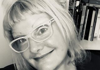 black and white portrait of Jane, head tilted, blond hair, glasses with white and grey striped frames, and a smile