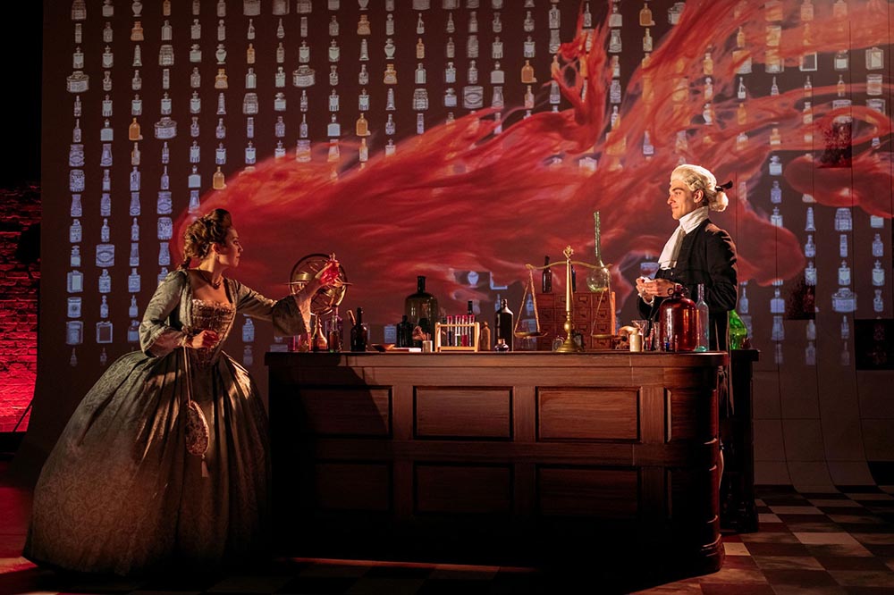 A man and a woman in period costume from around the 1600s by the look of the wig. They are standing either side of a chemists wooden counter and red smoke billows in the air