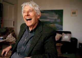 A photograph of Dannie Abse in a comfortable-looking study or sitting room.He is wearing a tweed jacket and a denim shirt, his hair is white and he is laughing. Photo credit Amit Lennon.