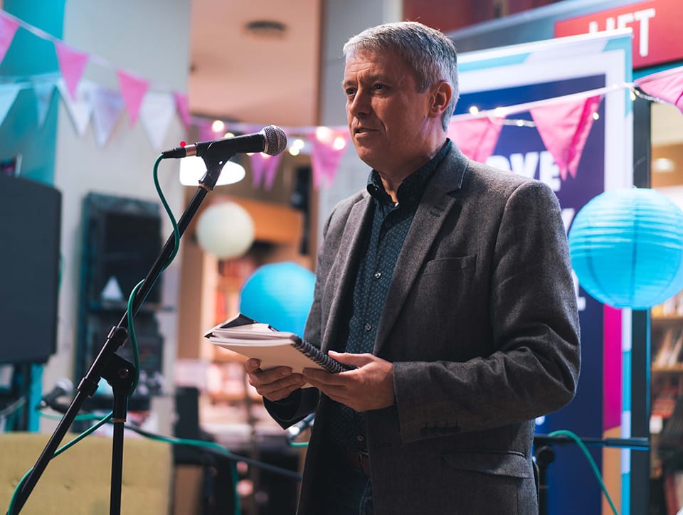 Johnathon Davidson reading into a microphone. He is wearing a grey suit jacket and a blue shirt and he has short grey hair