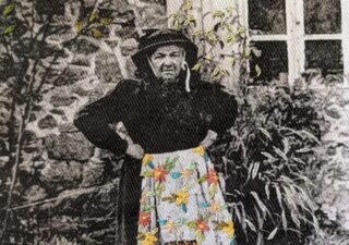 A predominantly black and white old photograph of an oldish woman standing if front of a stone house, arms akimbo. She is dressed in black and wearing a black bonnet-like hat. Her apron is in colour and embroidered with large primitive flower shapes.
