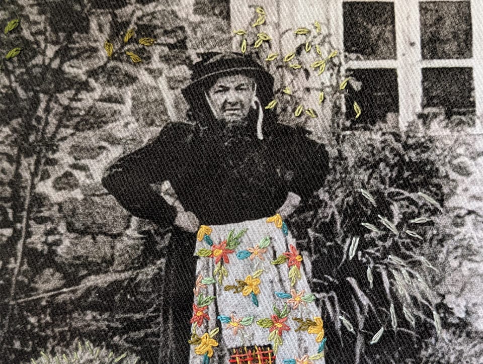A predominantly black and white old photograph of an oldish woman standing if front of a stone house, arms akimbo. She is dressed in black and wearing a black bonnet-like hat. Her apron is in colour and embroidered with large primitive flower shapes.
