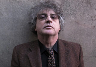 Paul Muldoon standing in front of a grey wall looking 'ard. He has grey curly hair and glasses, and he is wearing a brown tweed jacket and tie and a dark shirt.