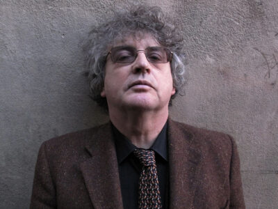 Paul Muldoon standing in front of a grey wall looking 'ard. He has grey curly hair and glasses, and he is wearing a brown tweed jacket and tie and a dark shirt.