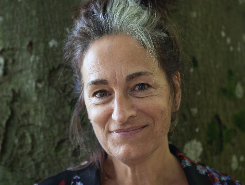 Greta Stoddart looking directly at the camera, smiling. She has brown hair with a grey streak and she's wearing a black shirt with small blue and red flowers on.