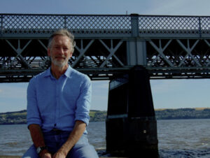 Photo of John Glenday sitting outside in front of a bridge. He is wearing a blue shirt and has a short grey beard and greying hair.