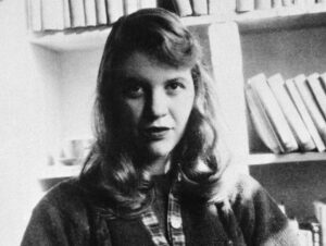 Black and white photograph of Sylvia Plath looking directly at the camera. There are bookshelves in the background. It can't be that warm because she's wearing a woolly cardigan with a plaid trim under the buttons.