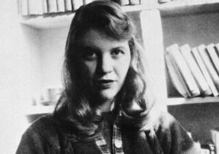 Black and white photograph of Sylvia Plath looking directly at the camera. There are bookshelves in the background. It can't be that warm because she's wearing a woolly cardigan with a plaid trim under the buttons.