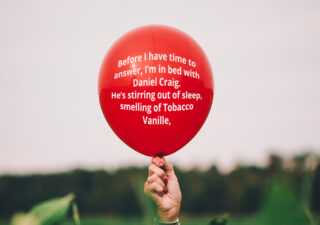 Hand holding a red balloon, written on the balloon in white text is the following "Before I have time to answer, I’m in bed with Daniel Craig. He’s stirring out of sleep, smelling of Tobacco Vanille."