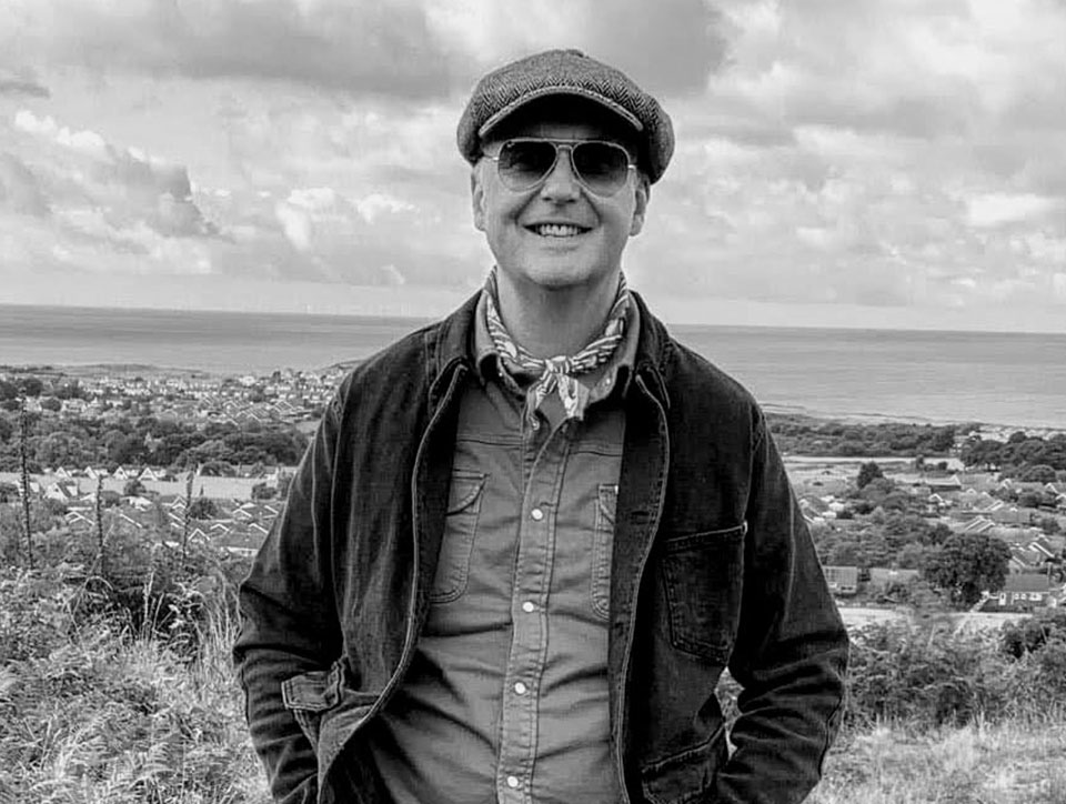 Black and white photo of Chris Hamilton-Emery. He is outside with sea in the background and is wearing a newsboy hat, tinted glasses and a neckerchief.