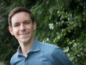Photo of Samuel Tongue. He is wearing a blue shirt and has short brown hair. He is smiling and standing in front of a green hedge.