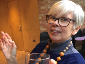 Photo of Kathy Pimlott. She has short grey hair, glasses and is holding a glass of wine. She is wearing ablue polo necked jumper and a necklace of orange wooden beads. Photo by Matthew Paul.