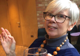 Photo of Kathy Pimlott. She has short grey hair, glasses and is holding a glass of wine. She is wearing ablue polo necked jumper and a necklace of orange wooden beads. Photo by Matthew Paul.