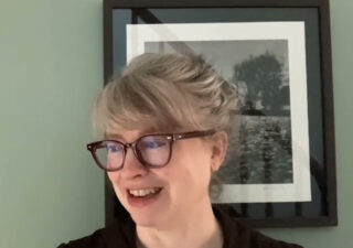 Natalie Shaw, wearing glasses and standing in front of a green wall with a framed painting on it.