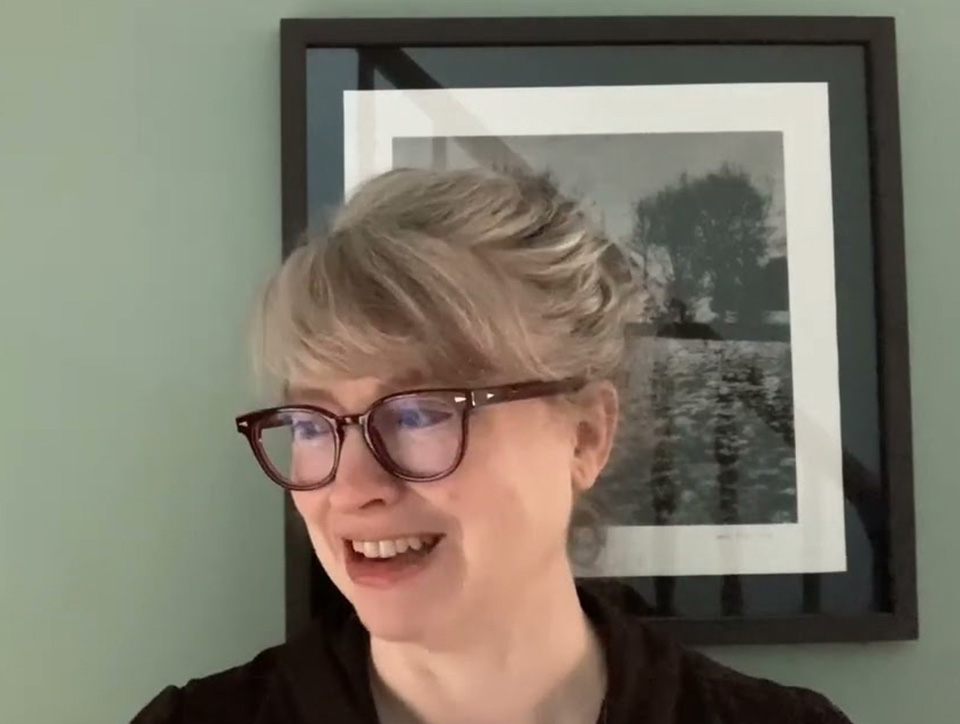 Natalie Shaw, wearing glasses and standing in front of a green wall with a framed painting on it.