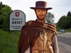 Clint Eastwood in full cowboy regalia standing in front of a road sign saying 'Welcome to Kent'.