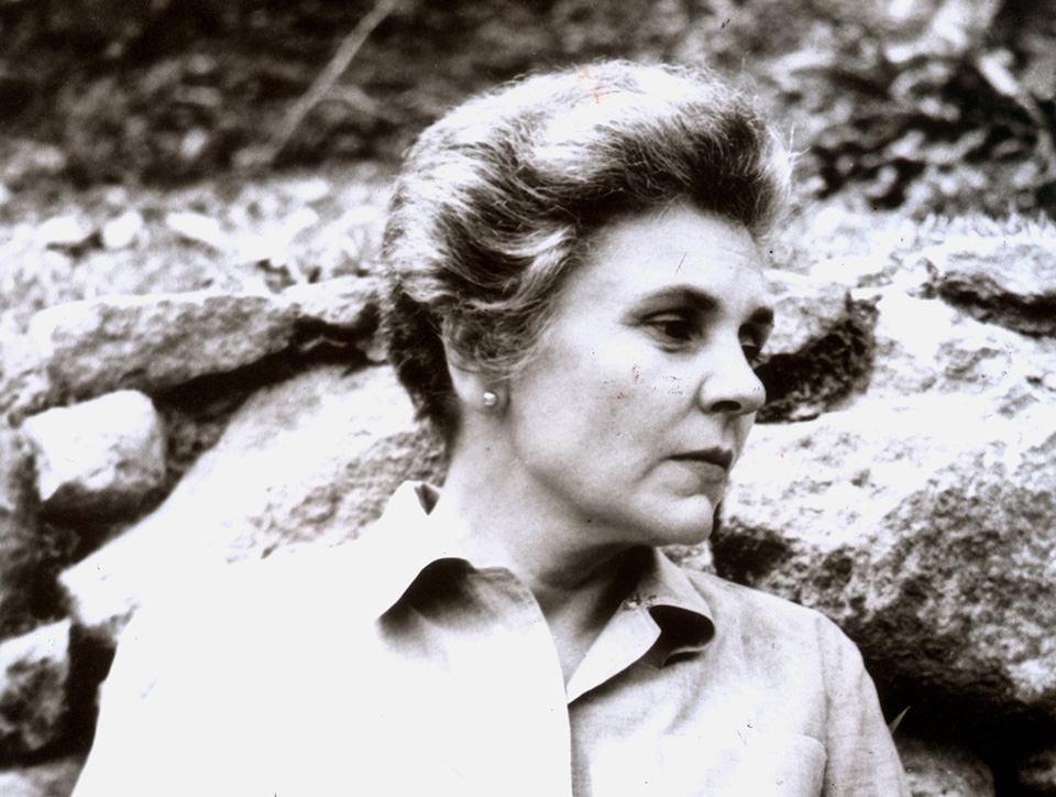 Black and white photo showing Elizabeth Bishop standing in front of some rocks.