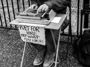 Black and white photograph showing a poet sitting outside at a tiny table with a typewriter. There is a handwritten sign saying "Poet for hire, pay what you like".