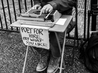 Black and white photograph showing a poet sitting outside at a tiny table with a typewriter. There is a handwritten sign saying "Poet for hire, pay what you like".