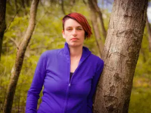 Photo of Kim Moore with a purple track suit style top. She has red hair and she is leaning against a tree.