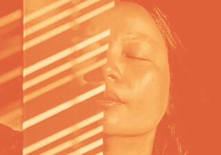 Mostly orange and orange tinted cover showing what looks like a woman's face half hidden by something that appears to be a vertical semi transparent column with thin diagonal white stripes on it.