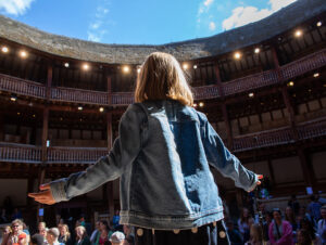 We see the back of a young girl standing on the stage at Shakespeare's Globe Theatre. She is wearing a denim jacket and her arms are outstretched.