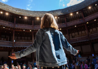 We see the back of a young girl standing on the stage at Shakespeare's Globe Theatre. She is wearing a denim jacket and her arms are outstretched.