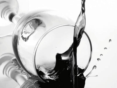 A close up black and white picture of a fallen wine glass, and dark liquid is splashing out.