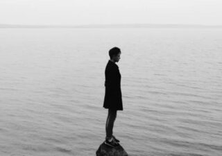 Monochrome photograph showing the silhouette of a woman standing on a small rock in the sea.