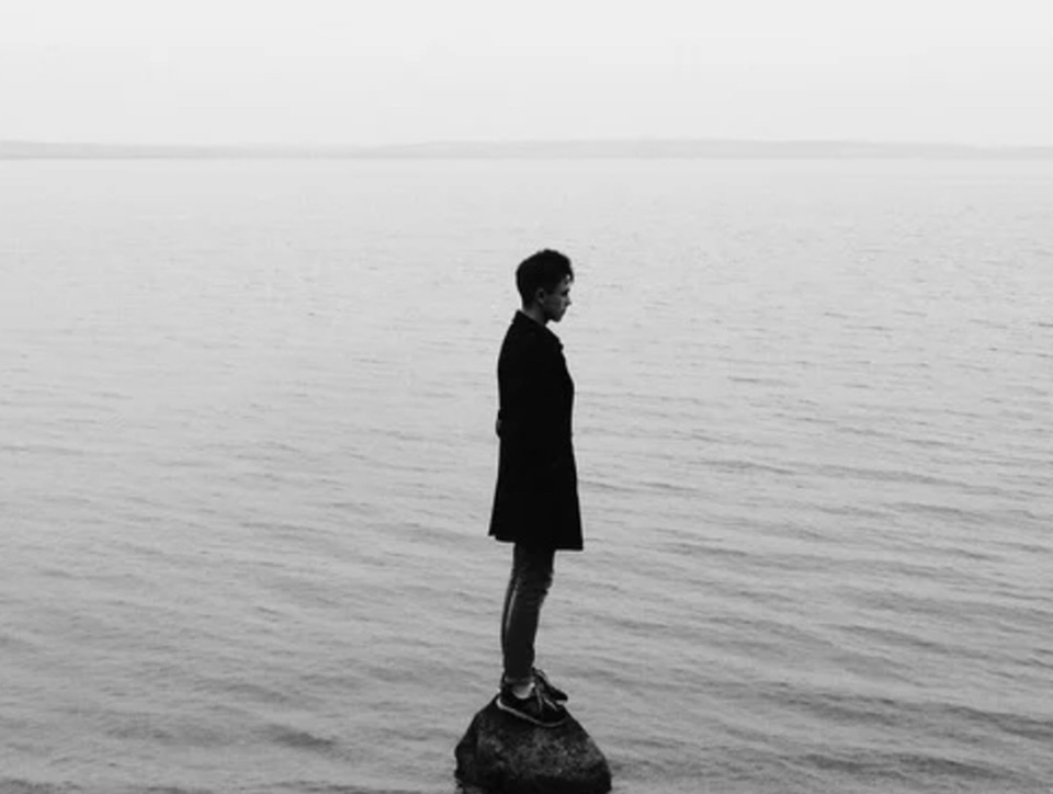 Monochrome photograph showing the silhouette of a woman standing on a small rock in the sea.