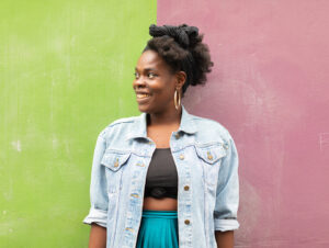 Photo of Vanessa Kisuule standing in front of a green and purple wall. She is looking to her right and smiling.