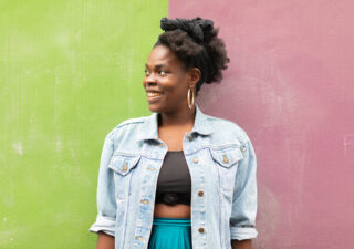 Photo of Vanessa Kisuule standing in front of a green and purple wall. She is looking to her right and smiling.